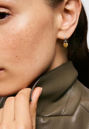 Gros plan d’une personne ajustant le col d’une veste en cuir olive foncé, portant une boucle d’oreille argentée en forme d’anneau avec un petit pendentif cœur en or.