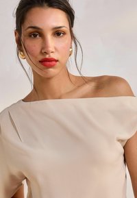 Vaatteena beige off-shoulder toppi, joka on valmistettu kevyestä kankaasta. Toppi on pehmeästi laskeutuva ja siinä on sileä pinta. Yksinkertaisia viivoja ja minimalistista suunnittelua korostava malli.