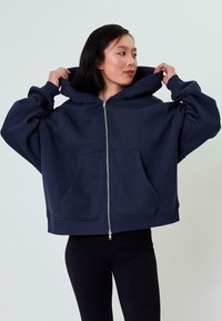 Felpa over-size blu navy con zip frontale, ampie tasche e cappuccio spazioso; realizzata in tessuto morbido e testurizzato.