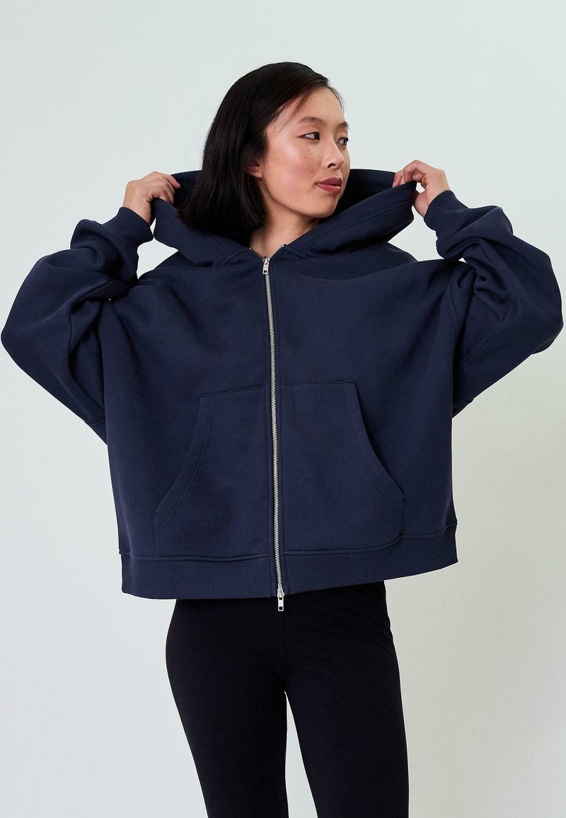 Felpa over-size blu navy con zip frontale, ampie tasche e cappuccio spazioso; realizzata in tessuto morbido e testurizzato.