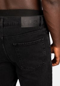 Närbild av en person som bär svarta jeans med en synlig bakficka och en sydd läderetikett som säger "Crystal Paris" på midjebandet.