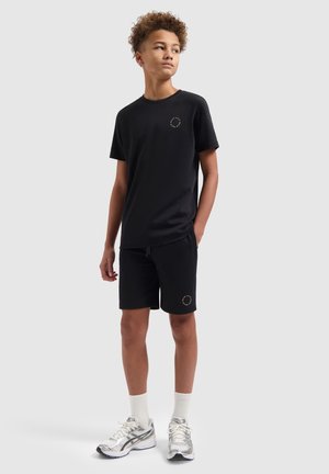 Tienerjongen staat, draagt een zwart T-shirt en korte broek met witte sportschoenen en sokken, kijkt iets naar links met één hand in de zak.