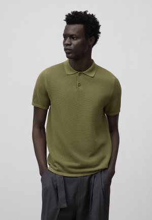 Polo shirt verde oliva in maglia testurizzata, con patta a tre bottoni e colletto classico, abbinato a pantaloni grigi a pieghe.