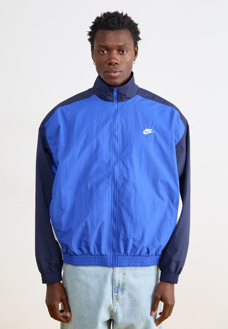 Nike vēja jaka ar zilu augšdaļu un tumši ziliem piedurknēm, ar rāvējslēdzēja priekšpusi, elastīgām aprocēm un mazu balto logo uz krūtīm.
