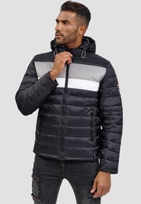 Chaqueta negra acolchada con rayas horizontales grises y blancas, cierre de cremallera, capucha ajustable y bolsillos laterales con cremalleras. Textura suave.