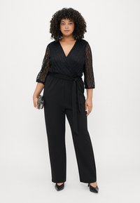 Svart jumpsuit med texturerade spetsärmar, omslagstyp och knytbälte. Har breda byxor och en subtil lyster. Levereras med en liten clutch.