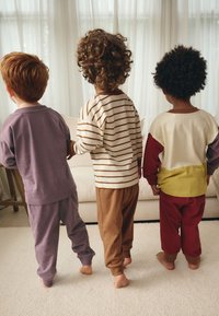 Trois enfants en vêtements d'intérieur : pantalons violet et marron, un t-shirt rayé crème, et un t-shirt à blocs de couleurs rouge, jaune et crème.