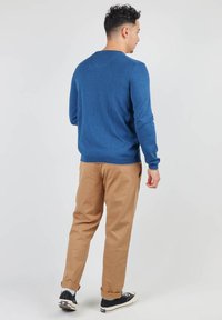 Pull en maille bleu, col rond, design ajusté. Associé à un pantalon chino beige et des baskets noires. Vue de dos, mettant en valeur la texture du pull.