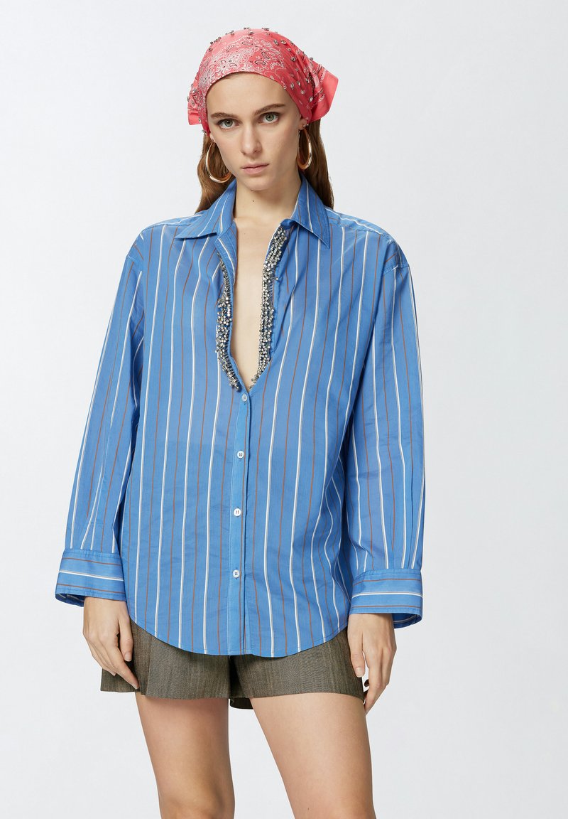 Camicia blu a righe con scollatura profonda decorata con embellishments argentati, abbinata a pantaloni corti grigi. Il modello indossa un foulard rosa con motivo.