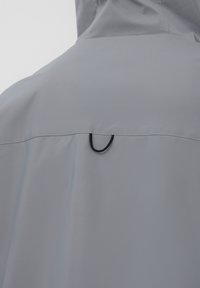 Vista posteriore di una giacca con cappuccio grigia che presenta una cucitura orizzontale con un piccolo anello nero nella parte superiore della schiena.