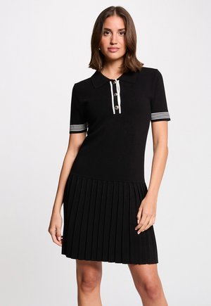 Kvinde iført en sort kortærmet kjole med hvidstribede kanter ved ærmerne og knappet krave, stående mod en ensartet lys baggrund.