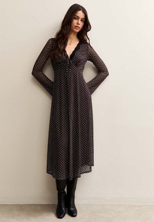 Woman wearing a black, long-sleeve, ankle-length polka dot dress with lace neckline, paired with black knee-high boots, standing against a wall.