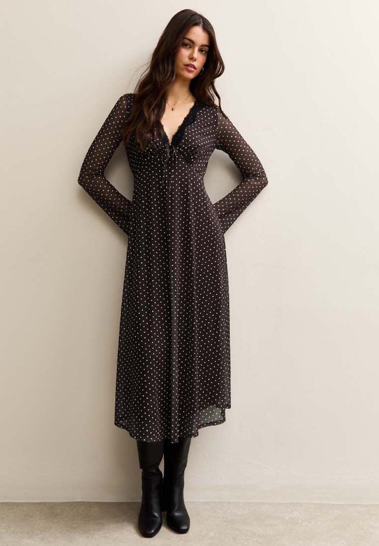 Femme portant une robe noire à manches longues, longueur cheville, à pois avec un col en dentelle, associée à des bottes noires montantes, se tenant contre un mur.