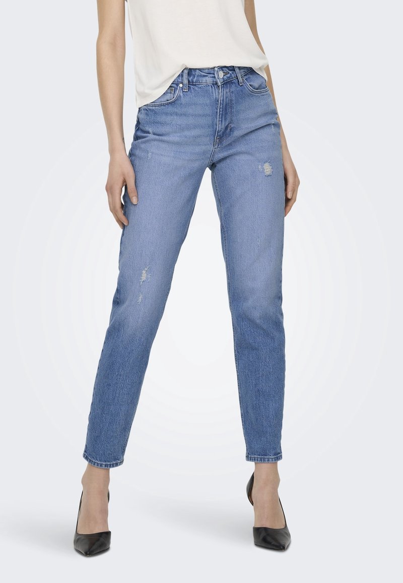 Femme portant un jean slim taille haute bleu clair avec légère usure et des escarpins noirs à bout pointu, debout devant un fond uni.