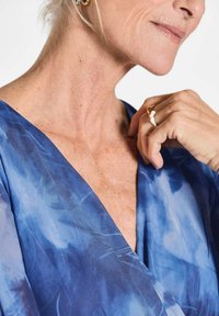 Blaues, fließendes Kleidungsstück mit tiefem V-Ausschnitt, das einen weichen, strukturierten Stoff mit einem subtilen abstrakten Muster präsentiert. Goldring-Akzent an der Hand.