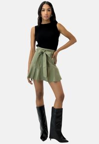Top negro sin mangas de canalé combinado con una falda verde oliva de talle alto, que presenta un lazo en la parte frontal y un dobladillo acampanado. Botas de tacón alto negras hasta la rodilla.