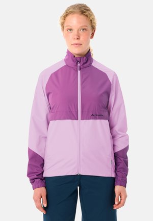 Jeune personne aux cheveux bouclés portant une veste zippée bicolore violette et un pantalon bleu foncé, debout devant un fond clair uni.