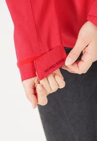 Röd skjorta med mjuk textur, utrustad med manschett och broderad etikett med texten "W-PRO 10000." Hand som greppar manschetten, vilket visar detaljer och passform.