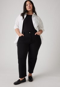 Jean en denim noir avec une coupe slim, associé à un t-shirt noir côtelé et une chemise blanche à boutons surdimensionnée. Des mocassins noirs complètent le look.