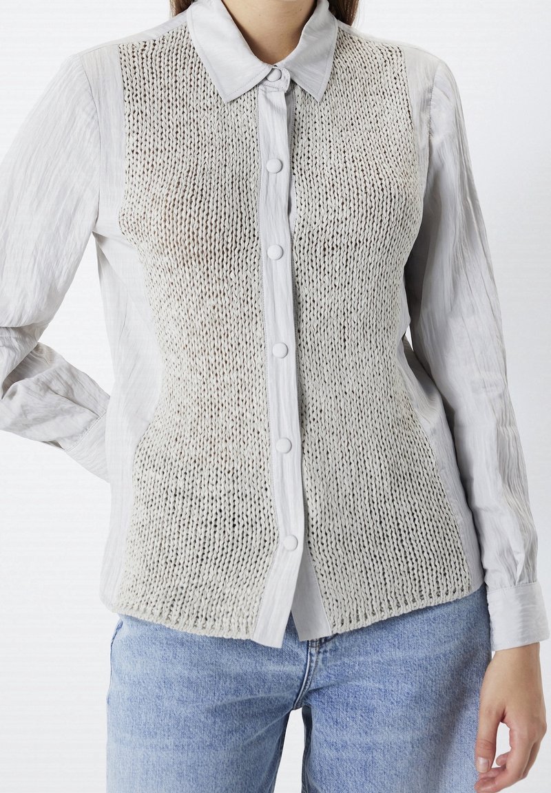 Femme portant une chemise à manches longues grise claire à boutons avec un gilet beige en tricot par-dessus et un jean bleu clair, debout devant un fond blanc.