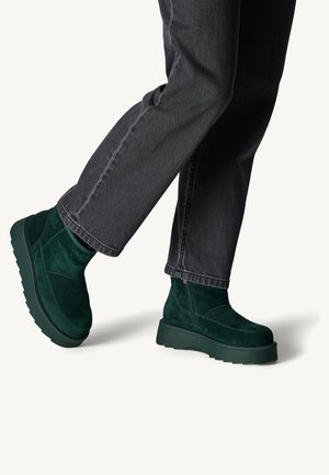 Green suede ankle boots with a thick platform sole, featuring stitching details and a smooth texture. Worn with dark grey jeans.