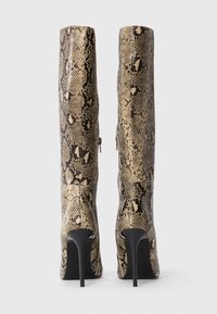 Un paio di stivali alti fino al ginocchio con tacchi a stiletto neri a punta e una trama in stampa serpente beige, mostrati da dietro su uno sfondo semplice.