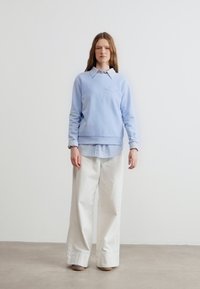 Γυναίκα με μακριά μαλλιά, που φοράει ανοιχτό μπλε πουλόβερ πάνω από πουκάμισο με γιακά και φαρδιά λευκή παντελόνα, στέκεται μπροστά σε έναν απλό λευκό τοίχο.