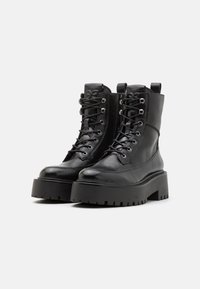 Botas de cuero negras hasta el tobillo con suelas de goma gruesas, diseño de cordones, punta redonda y costuras reforzadas. Incluyen una pestaña en la parte posterior.