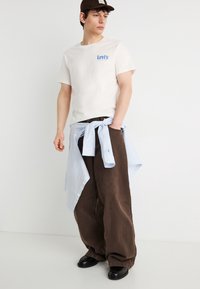 Νεαρός άνδρας που φοράει λευκό μπλουζάκι Levi's, καφέ φαρδιά παντελόνια, μαύρα παπούτσια και μπλε ριγέ πουκάμισο δεμένο γύρω από τη μέση, κοιτάζοντας προς τα πλάγια.