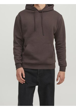 Mand iført en brun pullover-hoodie med forlomme og sorte bukser, stående mod en ensartet lys baggrund.