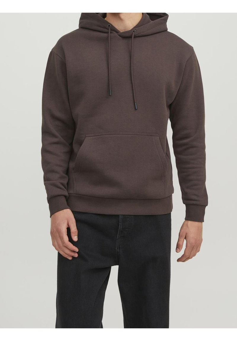 Homme portant un sweat à capuche marron avec poche avant et pantalon noir, debout devant un fond clair uni.