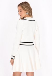 Weißes Strickkleid mit eng anliegendem Oberteil, ausgestelltem Rock, schwarzem Rand am Kragen und den Ärmeln, mit einem strukturierten Rautenmuster und einer definierten Taille.