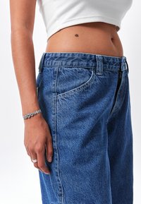 Jeans de mezclilla azul claro con cintura alta, que cuentan con bolsillos frontales y una cremallera. Se llevan con una blusa corta blanca y joyas de plata.