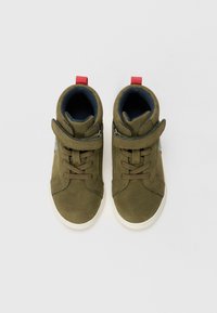 Baskets montantes vert olive en daim avec une semelle en caoutchouc blanc, dotées d'une sangle Velcro, d'un laçage et d'une languette rouge sur le talon.