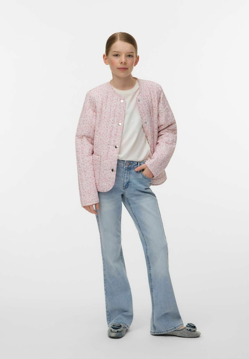 Noor isik seisab käsi taskus, kannab heleroosat mustrilist jopet, valget t-särki, hele siniseid teksapükse ja hallid paeltega kingi.