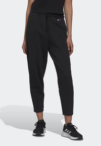 Žena v černých teplákových kalhotách Adidas s páskem na stažení a černých teniskách Adidas s bílou podrážkou, stojící na jednobarevném pozadí.