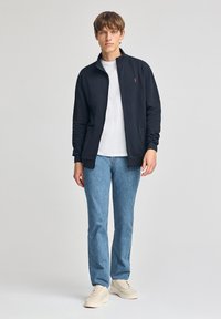 Giacca nera con zip realizzata in un materiale morbido, caratterizzata da un colletto alto e un piccolo logo ricamato, abbinata a jeans azzurri e sneakers beige.