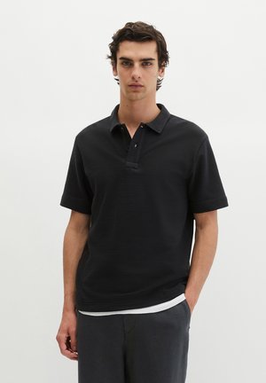Jonge man met donker haar die een zwart poloshirt met korte mouwen en donkere broek draagt, staand tegen een eenvoudige witte achtergrond.