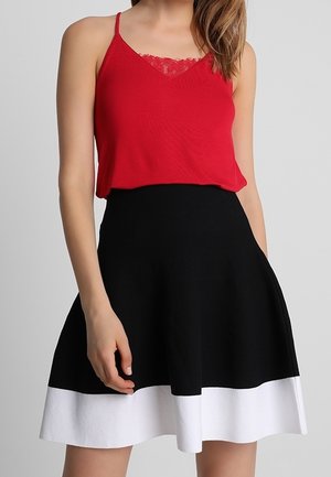 Femme portant un haut rouge sans manches avec un détail en dentelle et une jupe noire avec un large ourlet blanc, debout devant un fond gris.