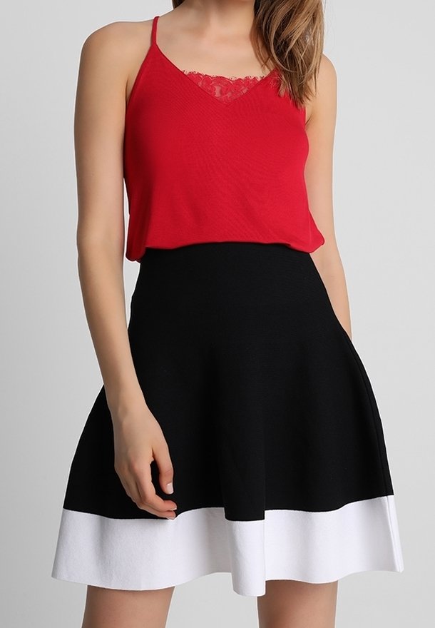 Femme portant un haut rouge sans manches avec un détail en dentelle et une jupe noire avec un large ourlet blanc, debout devant un fond gris.