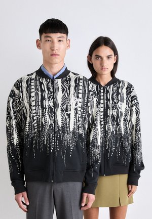 Giacca nera con zip, decorata con motivi astratti in bianco e grigio, dotata di colletto e polsini a costine, indossata da due modelli.