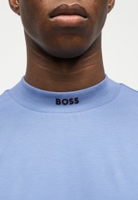 Κοντινό πλάνο ενός ατόμου που φοράει ανοιχτογάλανο μπλουζάκι με στρογγυλή λαιμόκοψη και τη λέξη "BOSS" με μαύρα γράμματα στο γιακά.