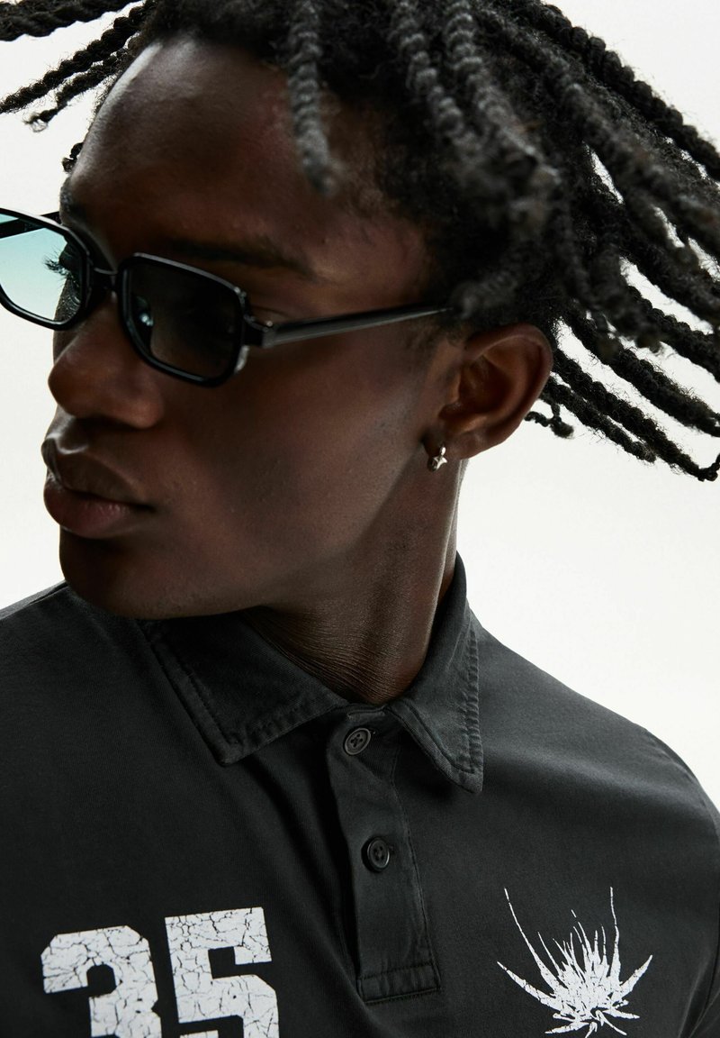 Sort polo skjorte med krave, der har et hvidt grafisk design og nummer "35." Stoffet ser glat ud, og knapperne er sorte.