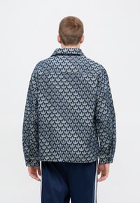 Temno modra jakna z vzorcem listov, sproščenega kroja in z ostrim ovratnikom. Narejena iz teksturirane tkanine z dolgimi rokavi in manšetami na gumb.