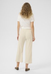 Damen kremfarbene weite Hosen aus strukturiertem Stoff mit elastischem Bund, kombiniert mit einem weißen T-Shirt und braunen Slippern.