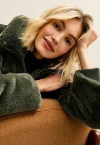 Mujer rubia con chaqueta peluda verde oscuro apoyada en una silla marrón, descansando la cabeza en una mano y sonriendo suavemente a la cámara.