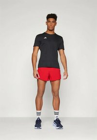 Crna sportska majica s logotipom, crvene kratke hlače i mornarsko plave tenisice za trčanje. Ima prugaste čarape i stojeći položaj. Jednostavan, funkcionalan dizajn.