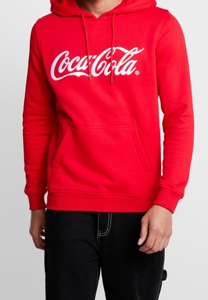 Person iført en rød hættetrøje med hvidt Coca-Cola-logo, forlomme og snore, kombineret med sorte bukser med synlige syninger.