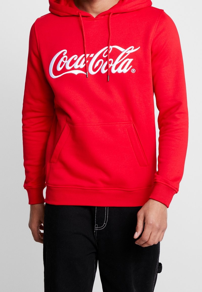Personne portant un sweat à capuche rouge avec logo Coca-Cola blanc, poche frontale et cordons, assorti d’un pantalon noir avec coutures apparentes.