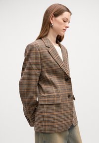 Blazer à carreaux marron avec de grands revers, deux boutons et poches avant. Texture lisse, présentant un motif à carreaux en beige et en teintes sombres.